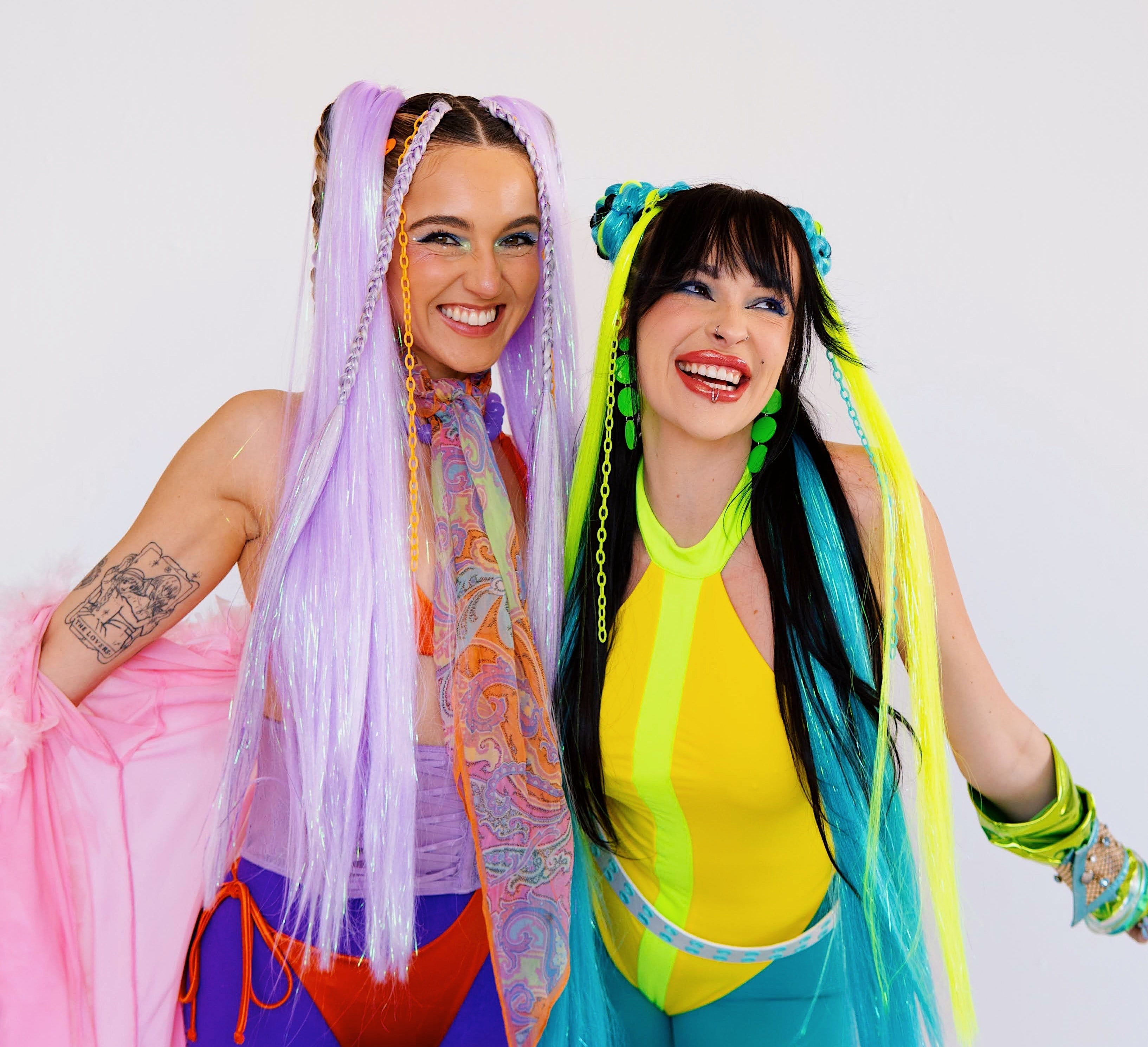 Two women with colorful hairstyles and outfits posing against a white background