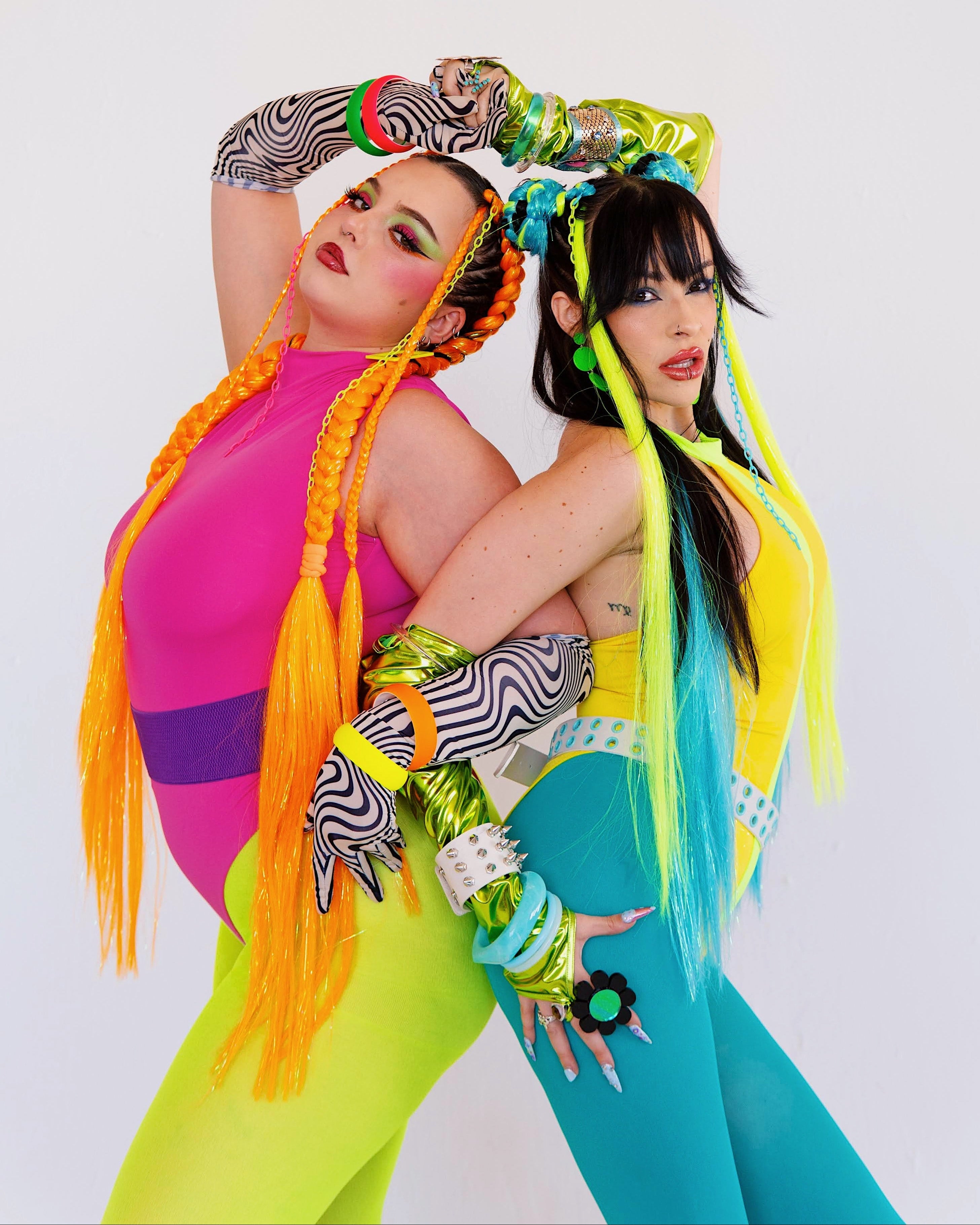 Two women in colorful outfits with headbands and hair extensions against a white background